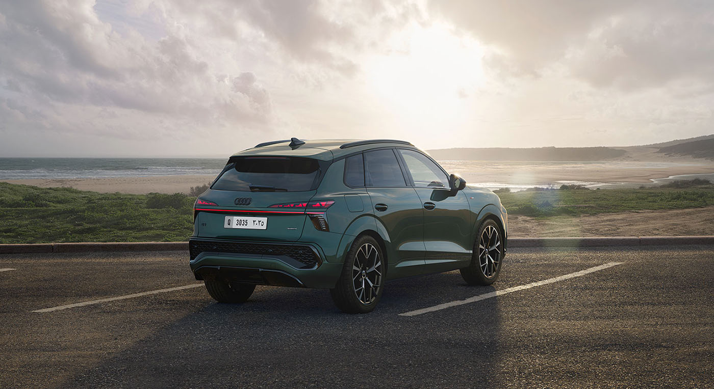 A sleek green Audi Q3 SUV is parked on a coastal road, facing away towards the ocean. The sun sets on the horizon, casting a warm glow on the scene.